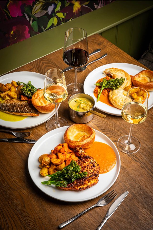 A Sunday roast spread at Vaasu Restaurant featuring spiced chicken, roasted fish, and a savory pie, each served with a Yorkshire pudding, vegetables, and wine.